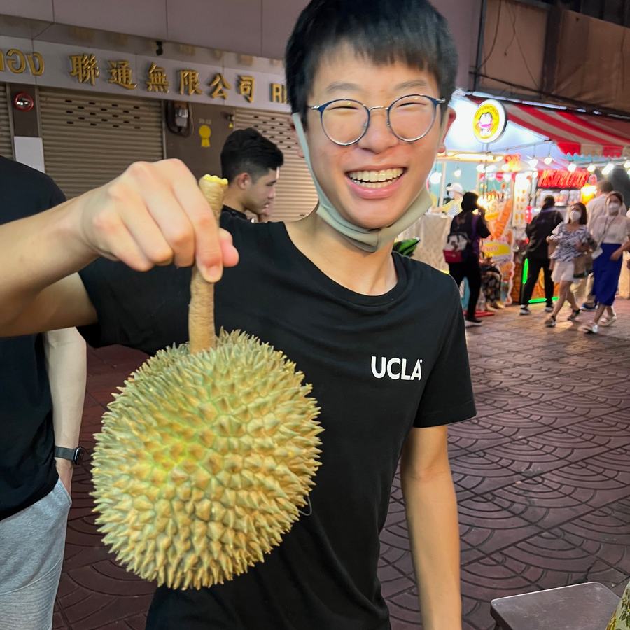 Ethan grinning and holding up a whole durian on a busy night street in Bangkok
