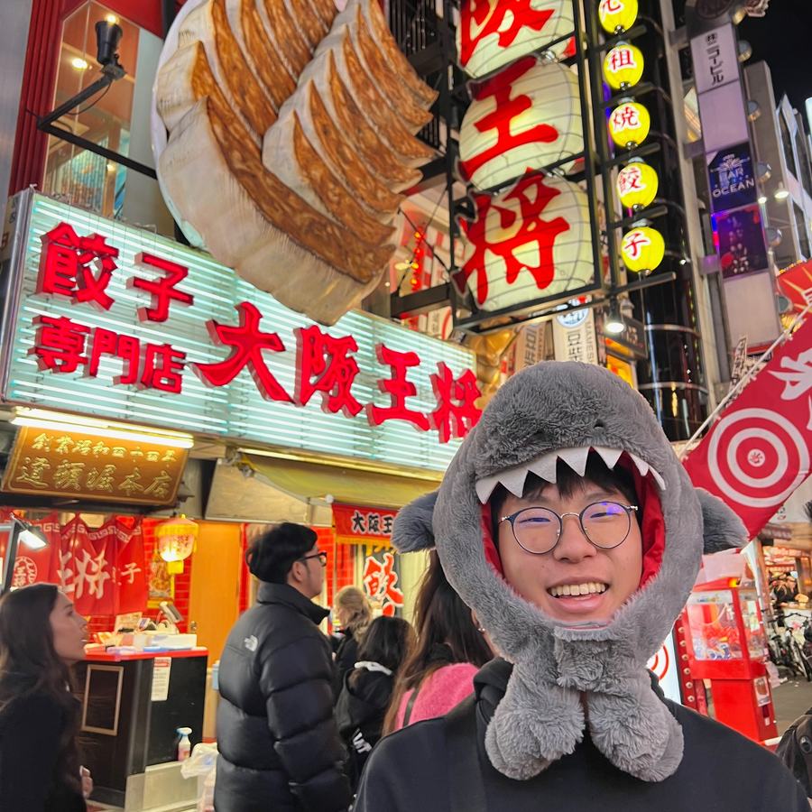 Ethan in a plush shark hat under the giant dumpling sign of a gyoza restaurant in Osaka