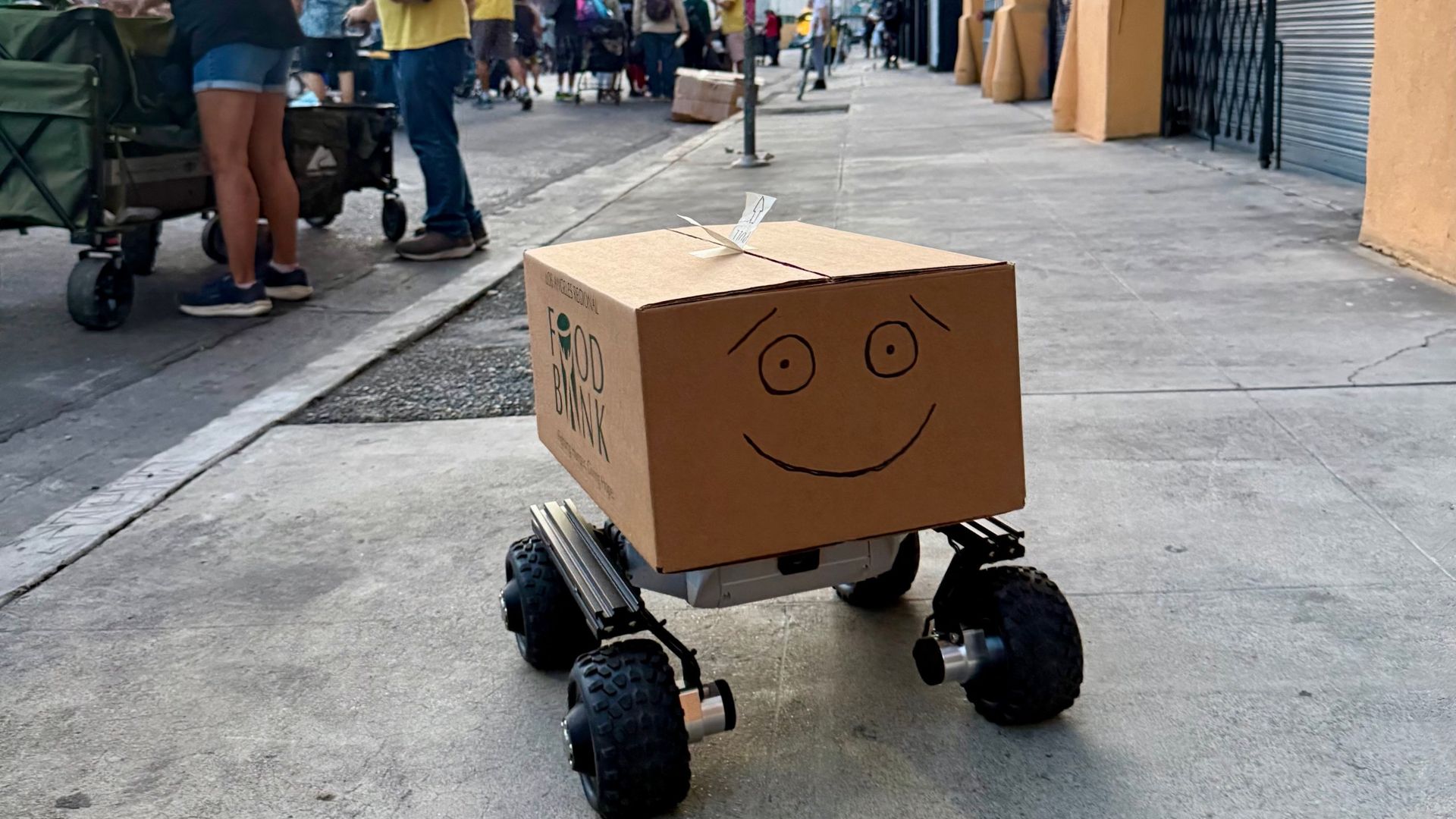 Food Angel, a cardboard box with a drawn smiley face on a wheeled robot base, on a sidewalk in Skid Row
