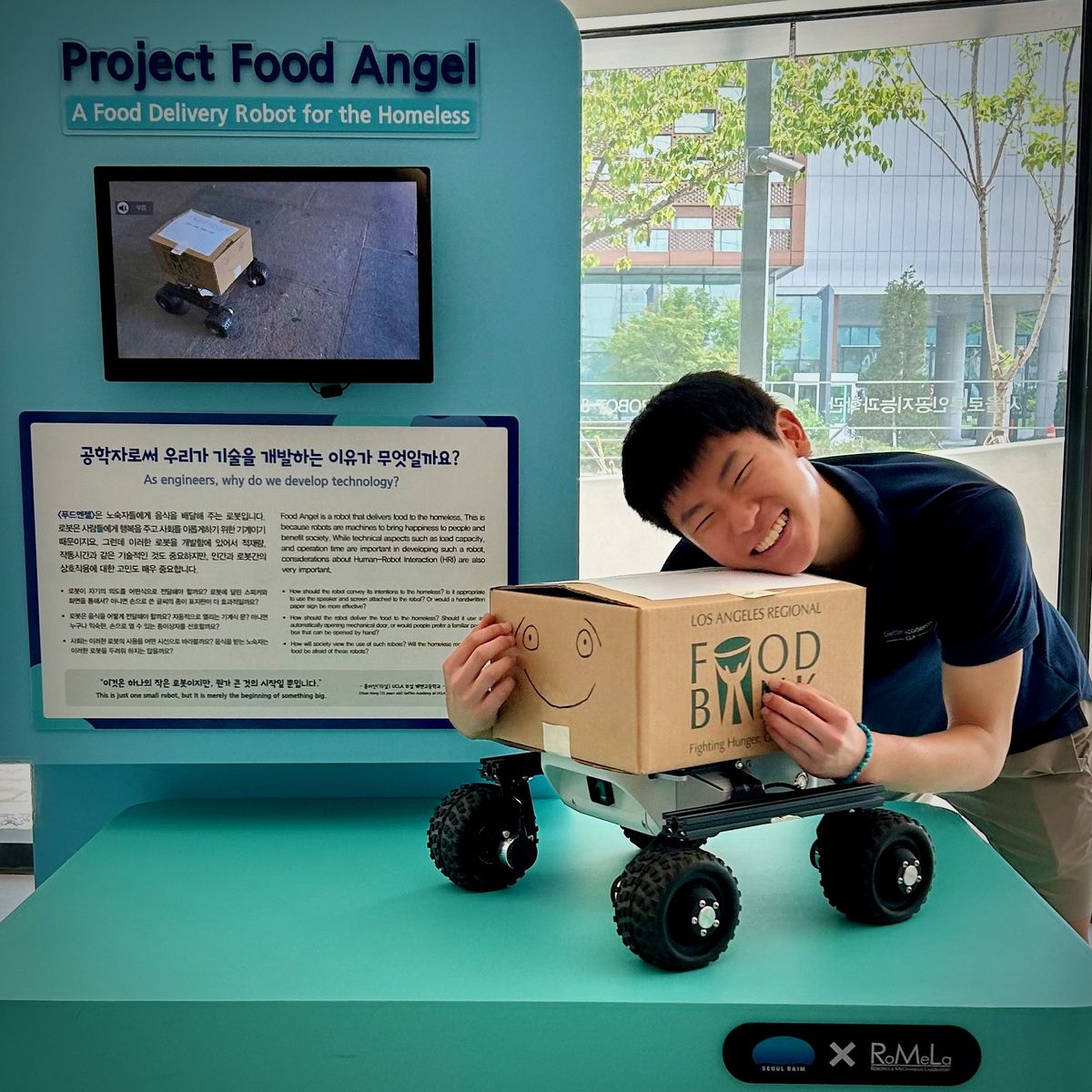 Ethan hugging Food Angel at its exhibit, under a sign reading Project Food Angel: A Food Delivery Robot for the Homeless