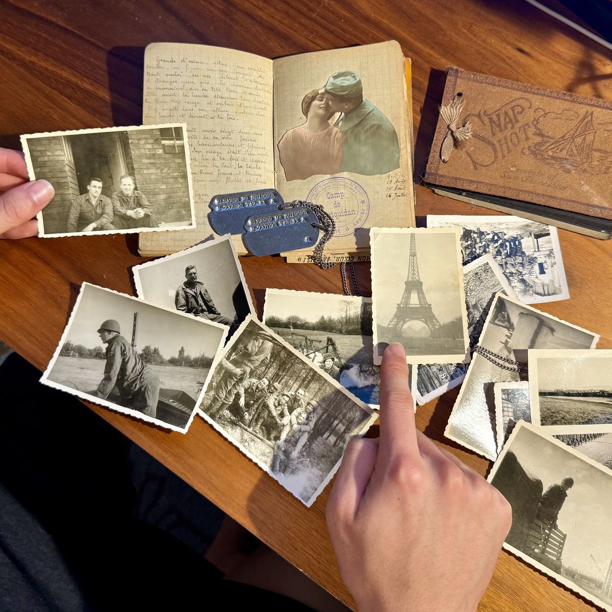 Ethan's hands sorting through old wartime black-and-white photographs, a handwritten diary, and dog tags on a table