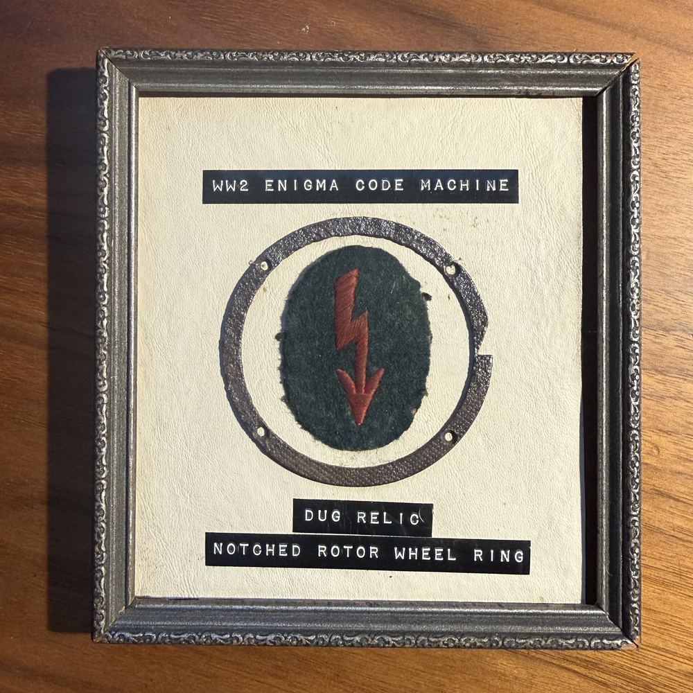 A framed relic labeled WW2 Enigma code machine, dug relic, notched rotor wheel ring