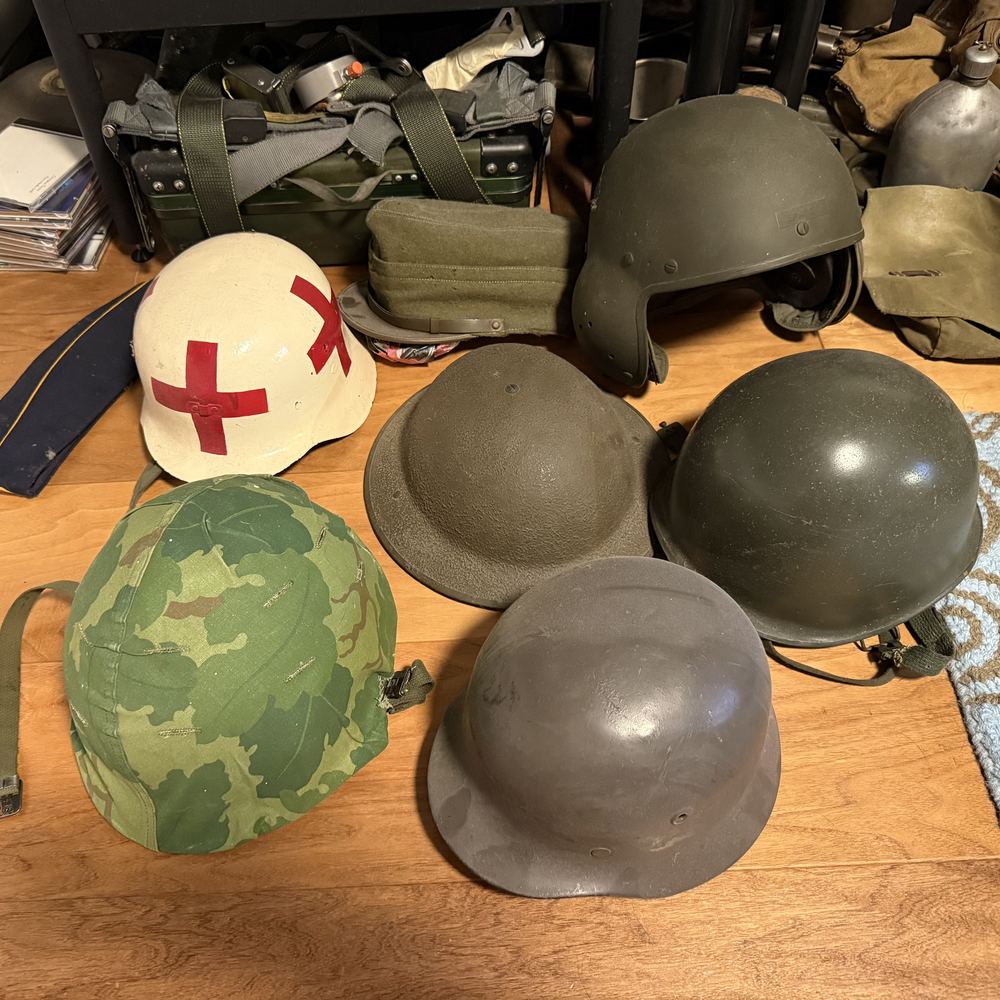 Seven military helmets on a table, including a white medic's helmet with a red cross, a camouflage-covered helmet, and a German-style steel helmet