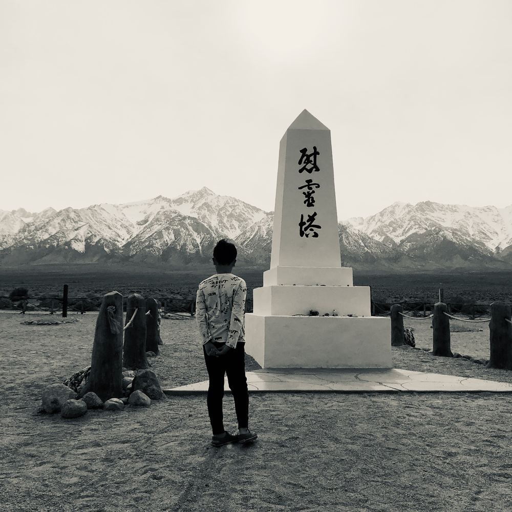 Young Ethan, seen from behind, standing before a white memorial obelisk with Japanese characters, with snow-covered mountains behind it
