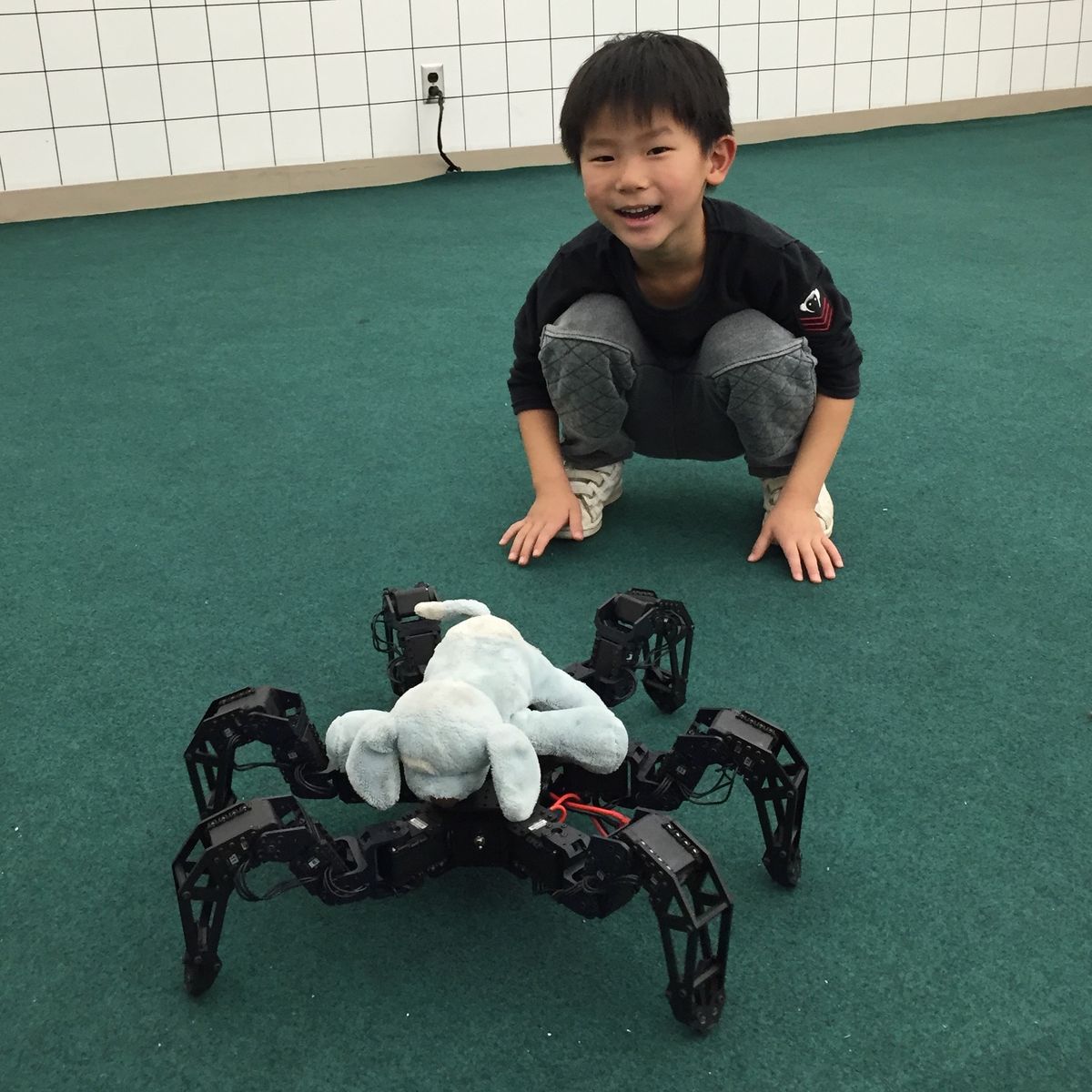Ethan as a young child crouching on a green carpet next to a six-legged robot carrying a stuffed animal