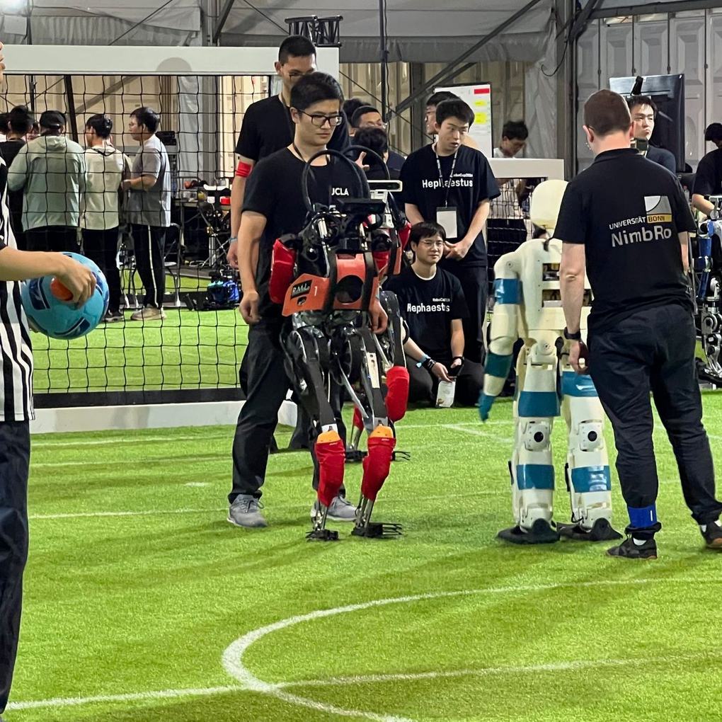 ARTEMIS walking onto the field beside a white-and-blue NimbRo humanoid at RoboCup 2024, with a referee holding the ball