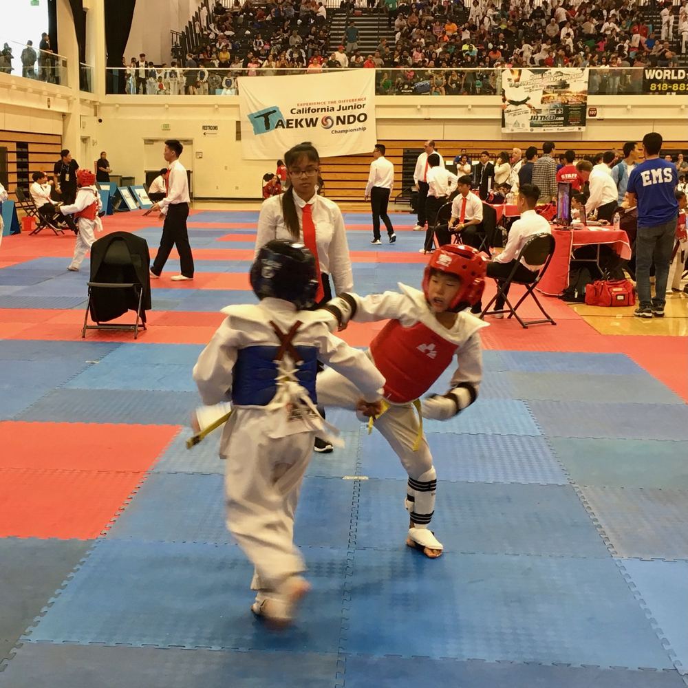 Young Ethan in a red helmet and chest protector kicking during a sparring match at the California Junior Taekwondo Championship